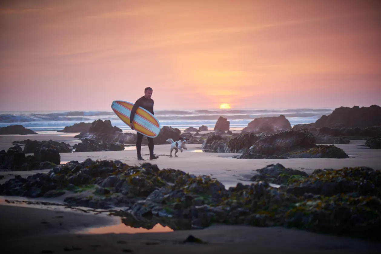 mature-surfer-leaving-the-surf-at-sunset-greeted-by-dog